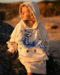 Kind sitzt auf einem Felsen am Strand in der Abendsonne im Ankerherz Hoodie EUROPA ONE LOVE Ivory Garment Dye, blauer Siebdruck Europa auf Zeus-Stier mit zwölf Sternen von Dani Garreton, Unisex Frontansicht