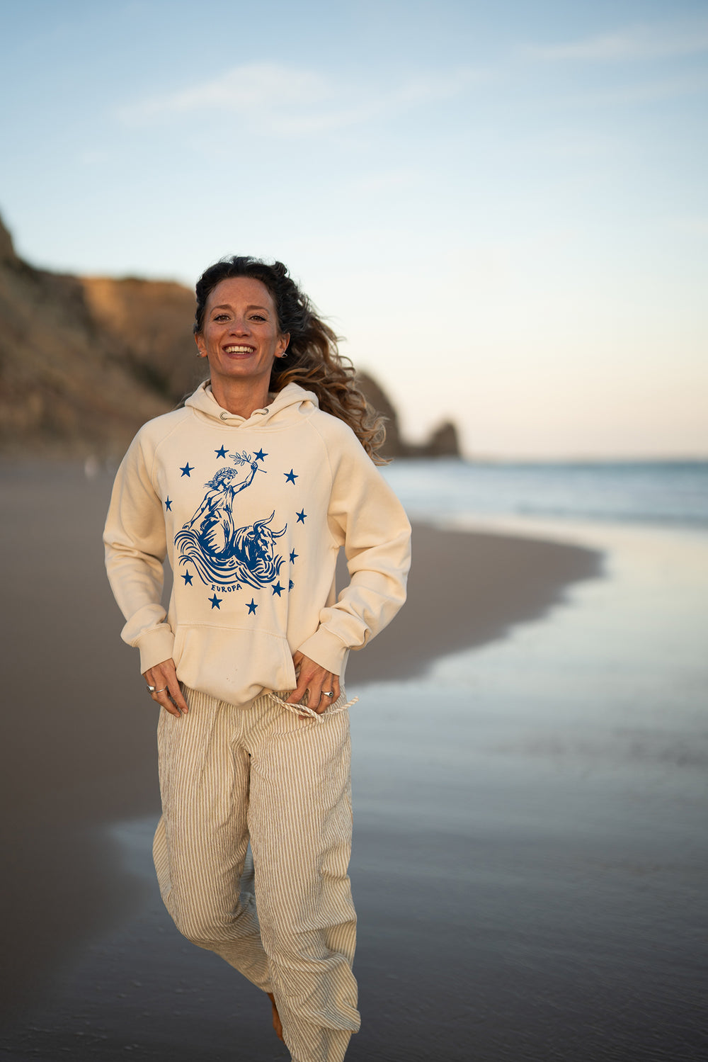 Frau läuft lachend über den Strand im Ankerherz Hoodie EUROPA ONE LOVE Ivory Garment Dye, blauer Siebdruck Europa auf Zeus-Stier mit zwölf Sternen von Dani Garreton, Unisex Frontansicht