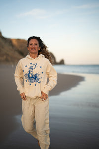 Frau läuft lachend über den Strand im Ankerherz Hoodie EUROPA ONE LOVE Ivory Garment Dye, blauer Siebdruck Europa auf Zeus-Stier mit zwölf Sternen von Dani Garreton, Unisex Frontansicht
